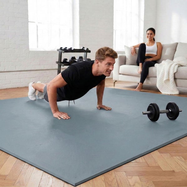Ein Mann macht Liegestütze auf einer grauen Bodenschutzmatte in einem hellen Fitnessraum mit Parkettboden und Hanteln.