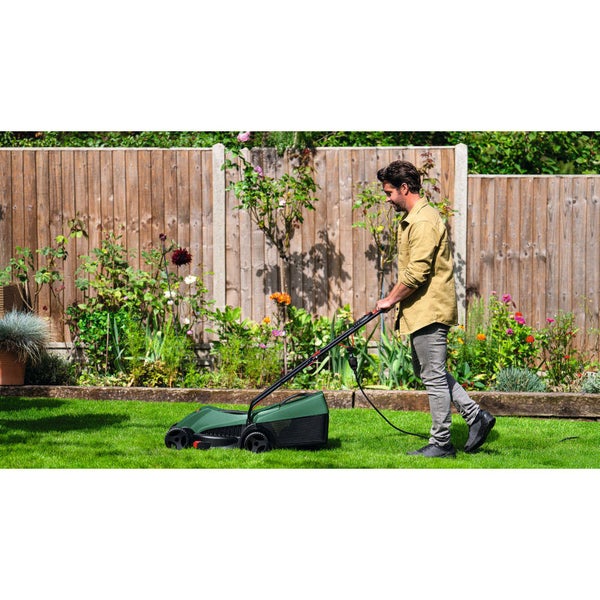 Ein Mann mäht einen grünen Rasen mit einem elektrischen Rasenmäher in einem hellen Garten mit Holzzaun und Blumenbeeten.
