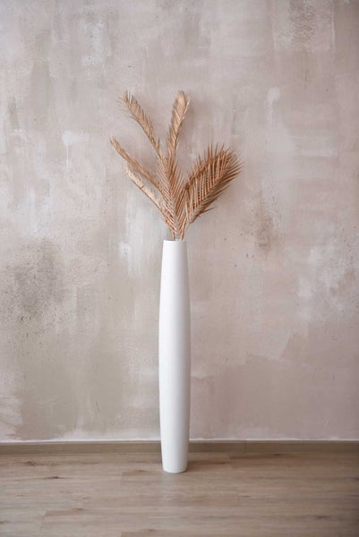 Hohe, schlanke Boden-Vase in Weiß mit dekorativen, goldfarbenen Palmwedeln vor einer Wand in Beton-Optik.