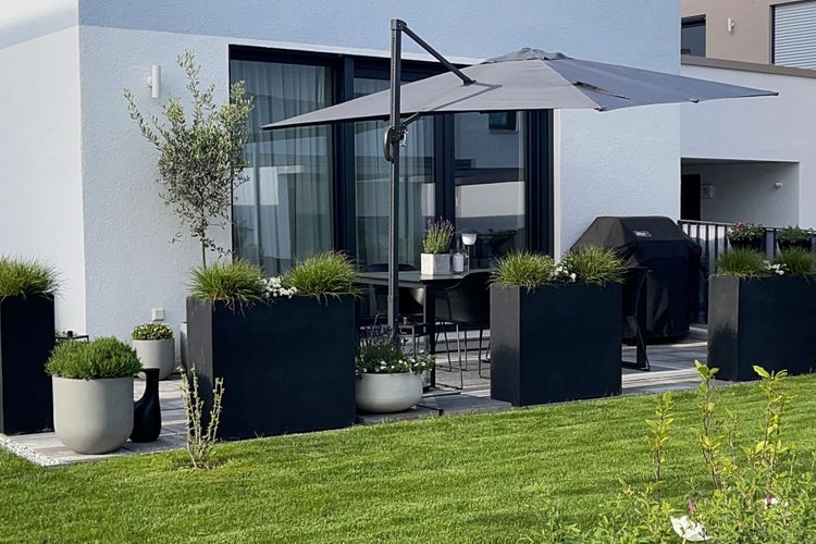 Moderne Terrasse mit grauem Ampelschirm, großen schwarzen rechteckigen Pflanzkübeln und Gartenmöbeln vor einer weißen Hauswand.