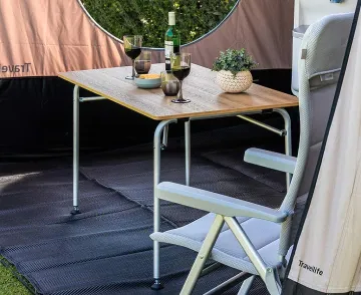 Campingtisch mit Tischplatte in Holzoptik und Metallgestell sowie ein grauer Campingstuhl mit Armlehnen in einem Vorzelt.