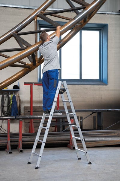 Handwerker steht auf einer Aluminium Stehleiter in einer Werkstatt und arbeitet an einer Metallkonstruktion.