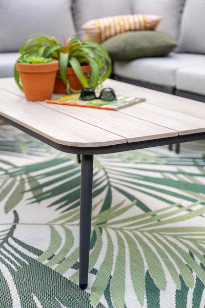 Moderner Garten-Couchtisch mit einer Tischplatte in Holzoptik und dunklem Metallgestell auf einem Outdoor-Teppich mit Palmenmuster.