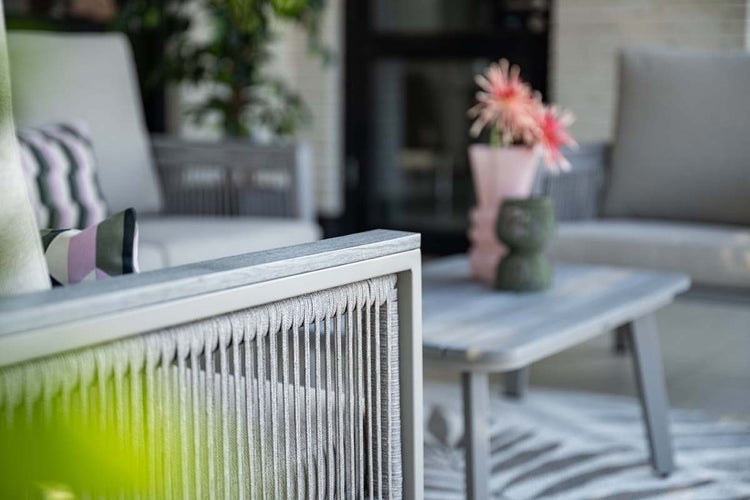 Nahaufnahme eines Garten-Lounge-Sessels mit Seilgeflecht und Holz-Armlehne, im Hintergrund ein Lounge-Set mit Couchtisch.