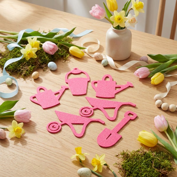 Filz Osterdekoration Set mit Gartenwerkzeug Motiven auf einem Holztisch mit Frühlingsblumen und Ostereiern arrangiert.