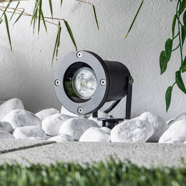 Schwarzer LED-Gartenstrahler platziert zwischen hellen Ziersteinen im Außenbereich vor einer Wand mit Grünpflanzen.