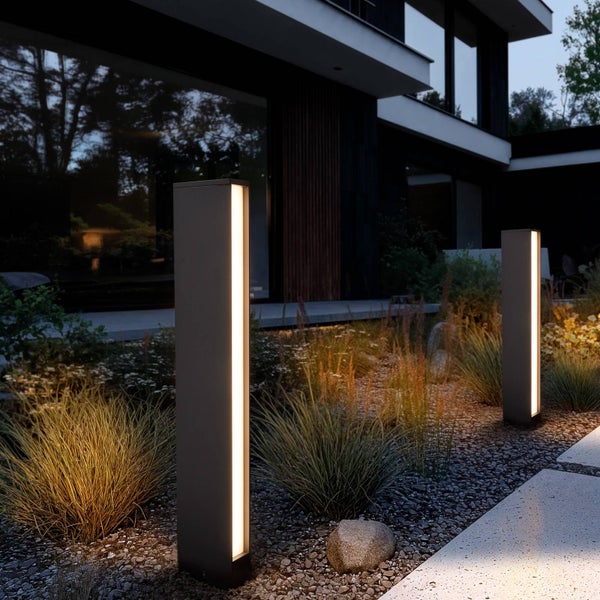 Moderne rechteckige Wegeleuchten beleuchten einen Gartenweg mit Kies und Ziergräsern vor einer modernen Hausfassade bei Nacht.