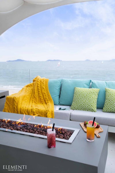Hell gestalteter Loungebereich auf einer Terrasse mit einem rechteckigen Gasfeuertisch, einem Sofa mit Textilkissen und Meerblick. Elementi Logo.