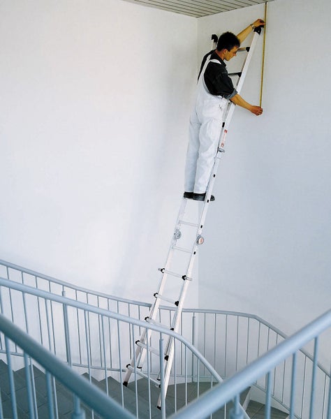 Ein Handwerker steht auf einer ausziehbaren Mehrzweckleiter im Treppenhaus und misst die Höhe einer Wand mit einem Maßband.