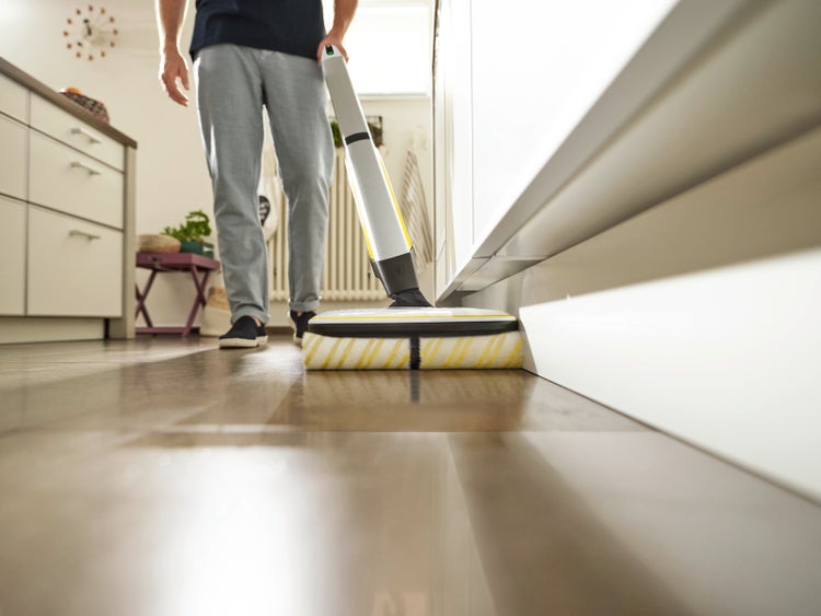 Frau reinigt mit einem Bodenreiniger unter einem Möbelstück auf einem Holzboden