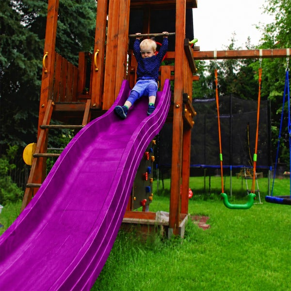 Kind auf einer lila Wellenrutsche an einem Spielturm aus Holz im Garten mit Schaukeln.