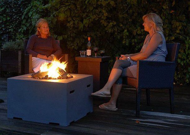 Zwei Personen sitzen abends auf einer Holzterrasse an einer quadratischen Gas-Feuerstelle in Betonoptik.