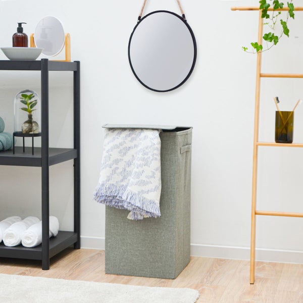 Faltbarer Wäschekorb aus grauem Stoff mit Deckel in einem hellen Badezimmer mit schwarzem Metallregal, rundem Spiegel und Holzleiter.