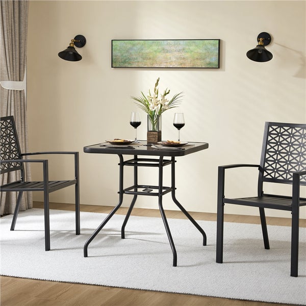 Sitzgruppe bestehend aus quadratischem Gartentisch mit Glasplatte und zwei schwarzen Metallstühlen mit geometrischem Muster in einem hellen Raum.