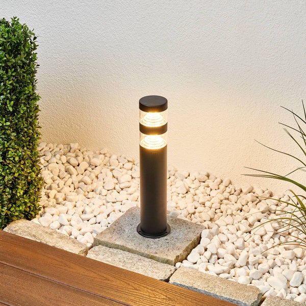 Moderne schwarze Pollerleuchte auf einem Steinfundament in einem weißen Kiesbeet, beleuchtet mit warmem Licht, neben einer Holzterrasse.