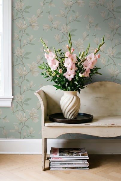 Vliestapete mit Magnolienblüten-Muster, Vintage-Sitzbank mit Blumenvase und Gladiolen vor gemusterter Wand.
