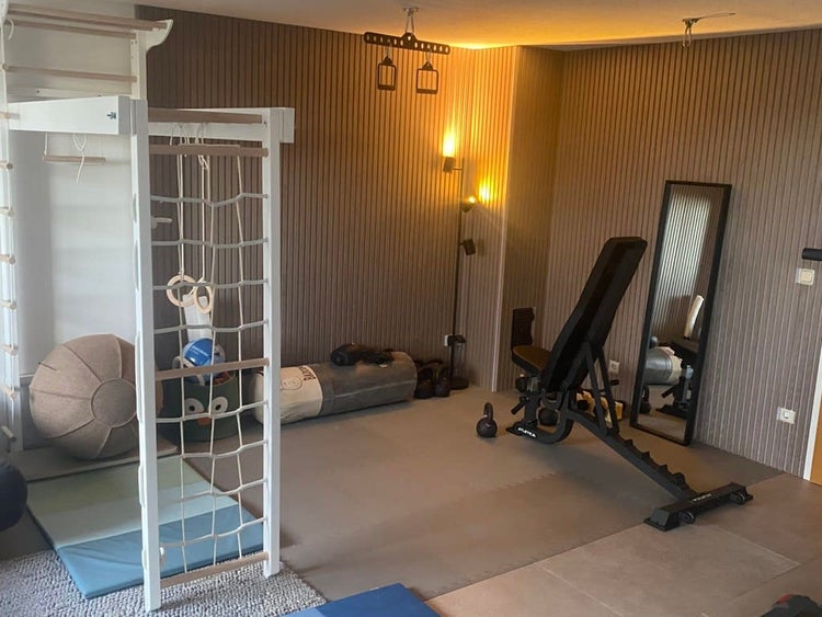 Heimfitnessraum mit Sprossenwand, verstellbarer Trainingsbank, Gymnastikball, Boxsack und Wandspiegel vor einer Lamellenwand.