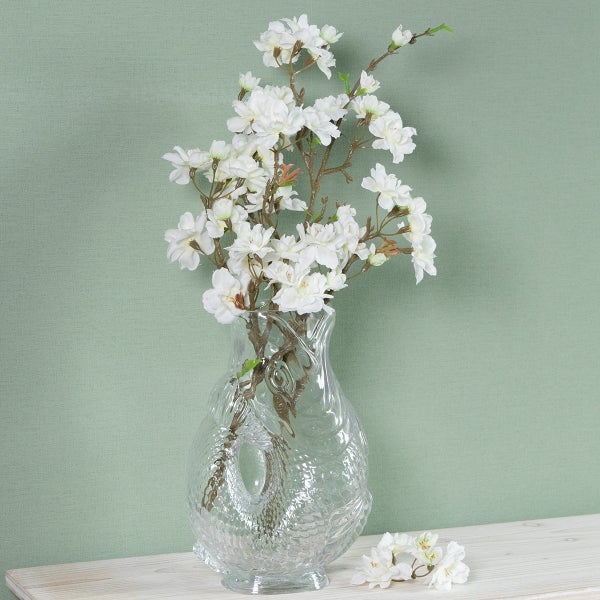 Künstlicher Kirschblütenzweig mit weißen Blüten in einer strukturierten Glasvase auf einem Holzregal.