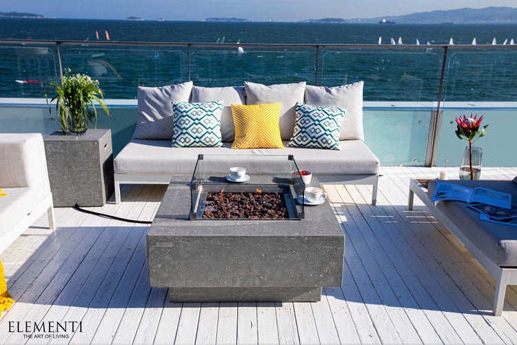 Quadratische Gas-Feuerstelle aus grauem Stein auf heller Holzterrasse, modern organisierte Lounge mit Sofa und Meerblick.