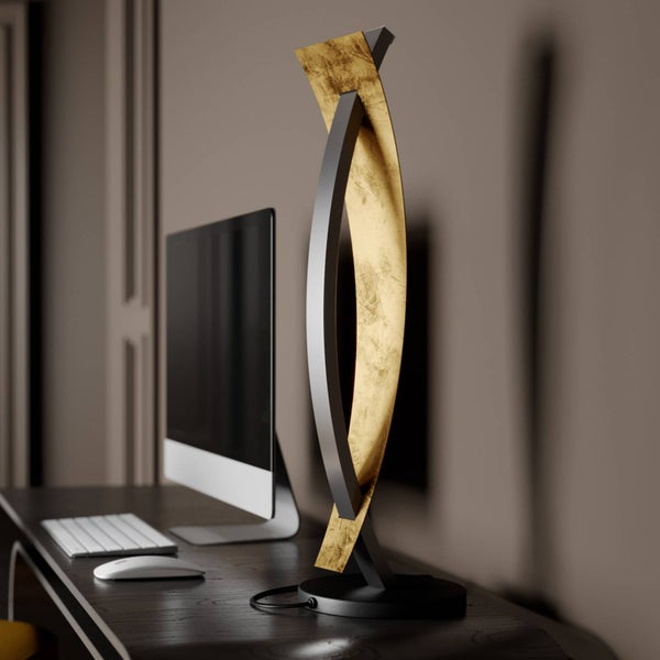 Moderne LED-Tischleuchte mit geschwungenem Design und Blattgold-Optik auf einem organisierten dunklen Holzschreibtisch neben einem Monitor.