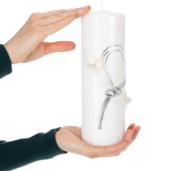 Weiße Stumpenkerze mit silberfarbenen Ringen, weißem Schleifenband und Herzdekor, gehalten von zwei Händen.