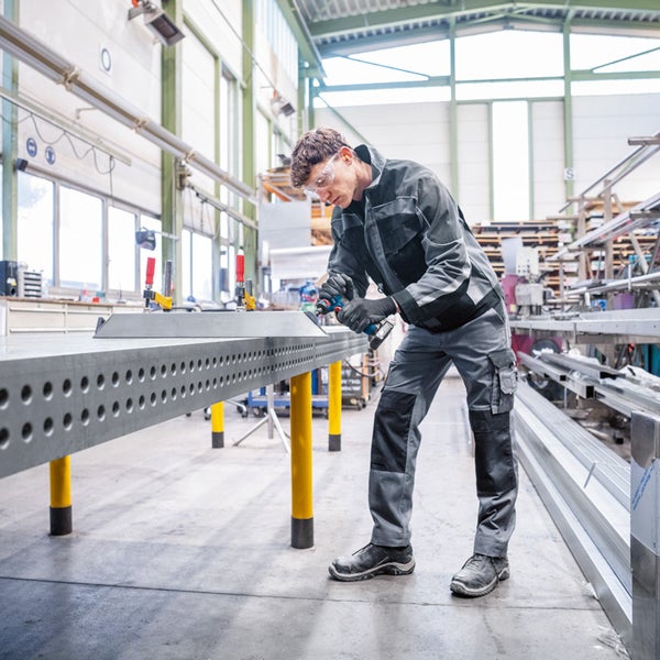 Ein Mann in Arbeitskleidung bearbeitet ein Werkstück mit einem Elektrowerkzeug an einem Werktisch in einer Werkshalle.