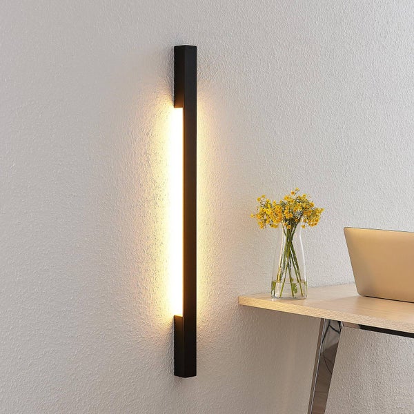 Moderne schwarze LED-Wandleuchte in vertikaler Form an einer Wand neben einem Schreibtisch mit Laptop und einer Vase mit gelben Blumen.