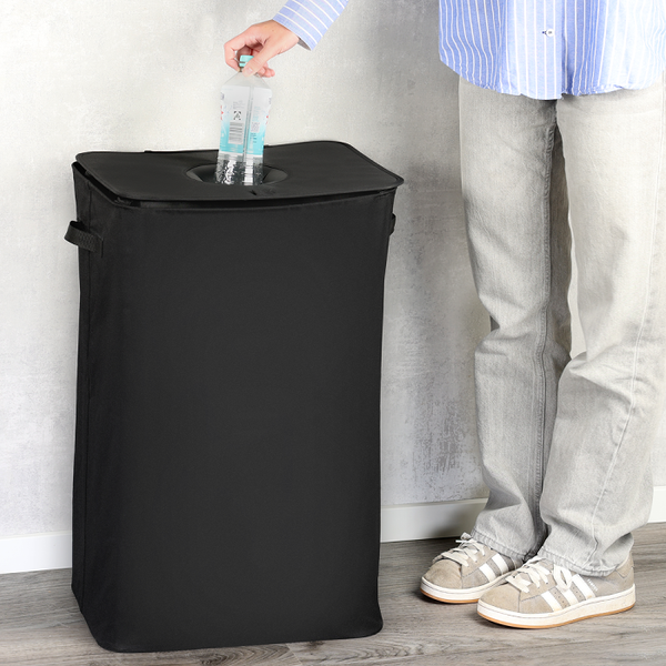 Wäschesammler oder Recyclingtasche aus schwarzem Stoff mit Deckel und runder Einwurföffnung, eine Person wirft eine Flasche ein.