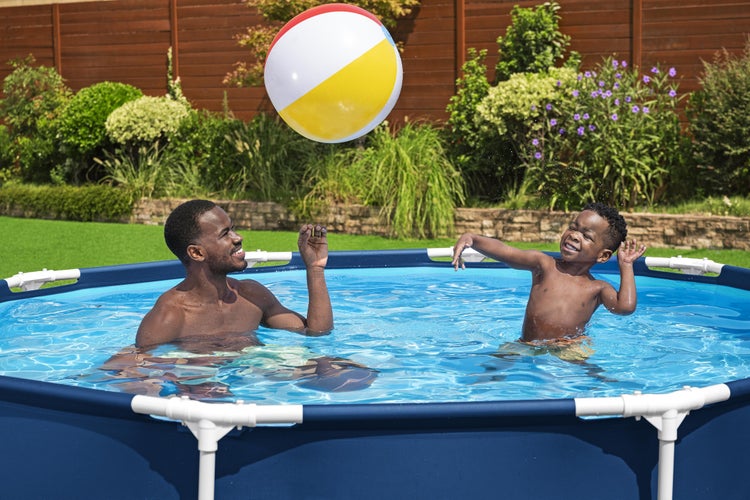 Ein Mann und ein Kind spielen mit einem Wasserball in einem runden Rahmenpool in einem hellen Garten mit Pflanzen und einem Holzzaun.
