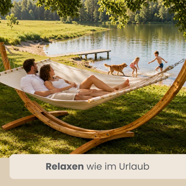Ein Paar entspannt in einer Stoffhängematte mit Holzgestell auf einer Wiese am See. Kinder und ein Hund spielen im Hintergrund.