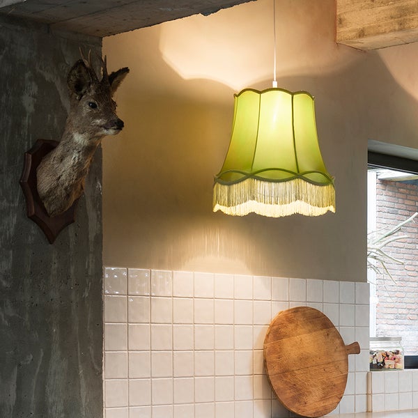 Grüne Pendelleuchte mit Stoffschirm und Fransen in einer Küche mit weißen Fliesen, einem Holzschneidebrett und Wanddekoration.