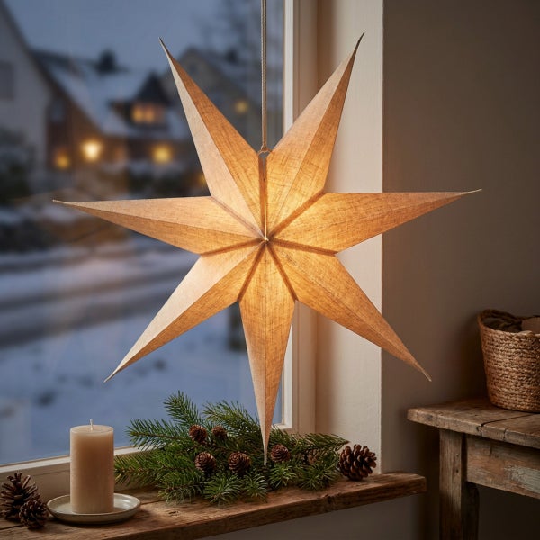 Beleuchteter Weihnachtsstern aus Stoff haengend vor einem Fenster dekoriert mit Tannenzweigen und Kerze auf einer Holzfensterbank.