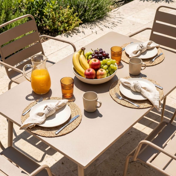Moderner, organisierter Gartentisch aus Metall mit Stühlen auf einer hellen Terrasse, gedeckt mit Geschirr, einer Obstschale und einer Saftkanne.
