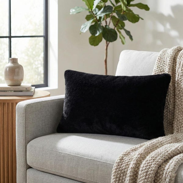 Schwarzes, rechteckiges Dekokissen aus flauschigem Material auf einem hellen Sessel in einem hellen Wohnzimmer.