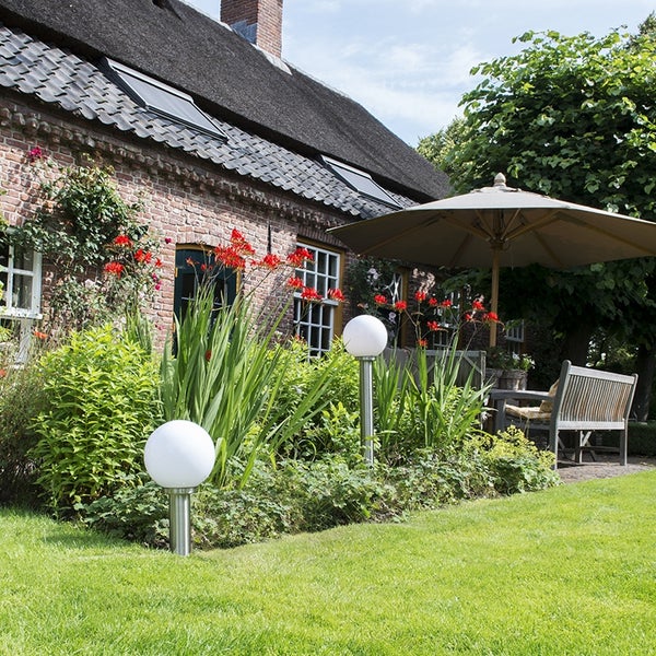 Zwei weiße kugelförmige Außenleuchten auf Edelstahlpfosten in einem Gartenbeet neben einem Rasen vor einem Haus mit Reetdach.