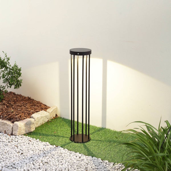 Moderne schwarze Außen-Sockelleuchte im zylindrischen Käfig-Design auf Kunstrasen, die eine Wand im Garten beleuchtet.