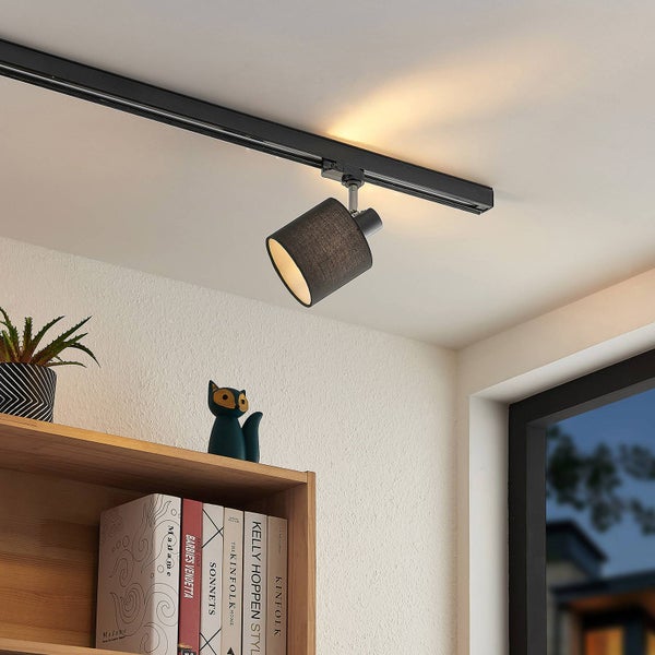 Schwarzer Schienensystem-Strahler mit Stoffschirm, montiert an einer Deckenschiene über einem Bücherregal aus Holz in einem hellen, modernen Raum.