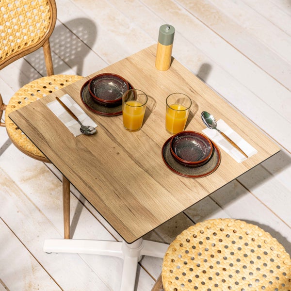 Quadratischer Gartentisch in Holzoptik mit Rattan-Stühlen und gedecktem Frühstücksgeschirr auf einer hellen Holzterrasse.