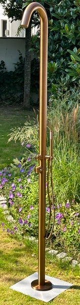 Moderne Gartendusche aus gebürstetem Metall in Kupferoptik mit Kopf- und Handbrause, aufgestellt in einem Garten mit Pflanzen und Blumen.