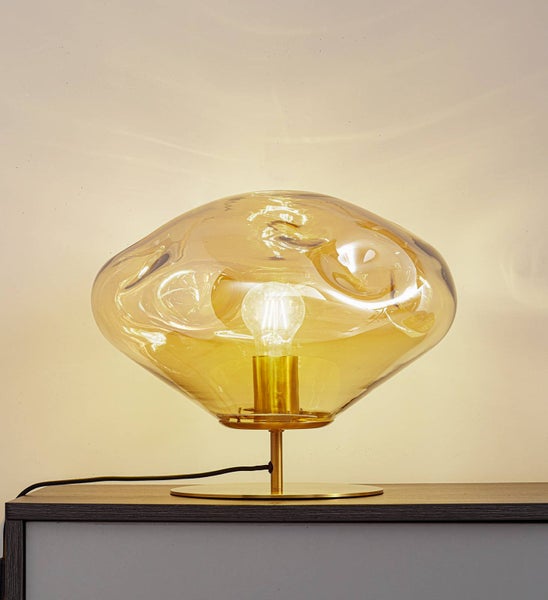 Tischleuchte mit unregelmäßig geformtem Glasschirm in Goldoptik und rundem Metallfuß, beleuchtet mit sichtbarem Leuchtmittel.