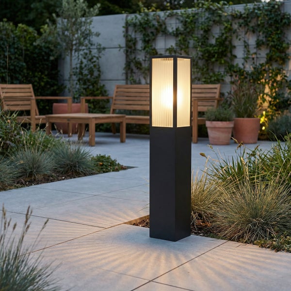 Moderne dunkelgraue Pollerleuchte mit geriffeltem Glas beleuchtet einen Gartenweg mit Steinplatten, Holzmöbeln und Gräsern.