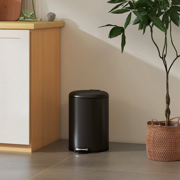 Schwarzer Treteimer in einem hellen, modern eingerichteten Raum neben einem Holzschrank und einer Pflanze im Korb.