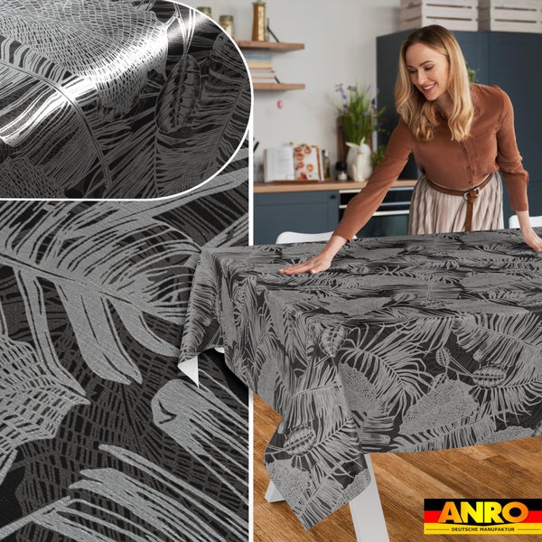 Eine Frau breitet eine Tischdecke mit botanischem Blattmuster auf einem Tisch in einer hellen Küche aus. Detailansicht der Textur. ANRO Logo.