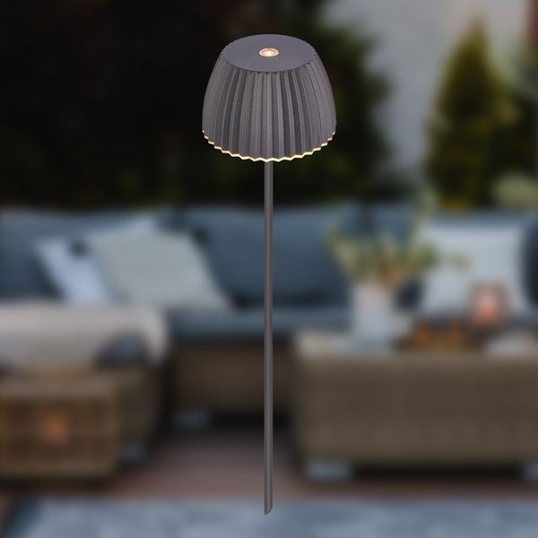 Solar LED Gartenleuchte mit gefaltetem anthrazitfarbenem Schirm und Erdspieß vor einem unscharfen Terrassenhintergrund.