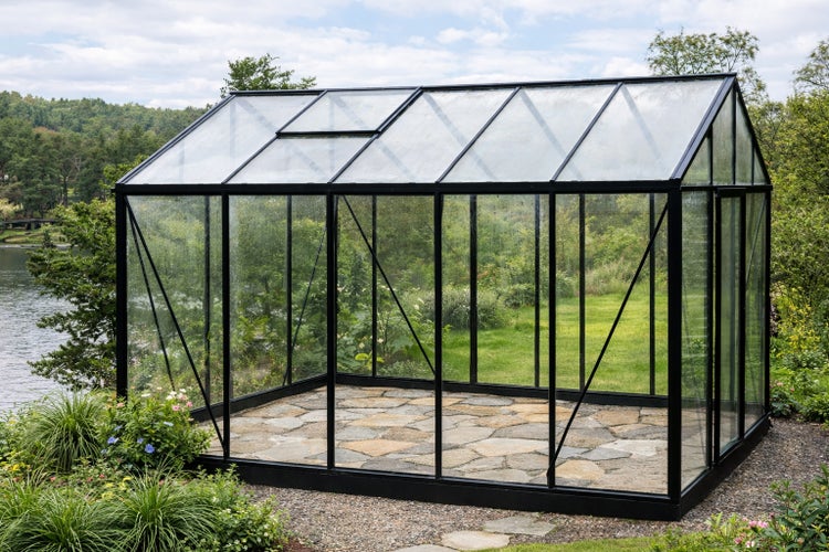 Modernes Gewächshaus mit schwarzem Metallrahmen und Glaswänden auf Steinboden in einem Garten am Wasser.