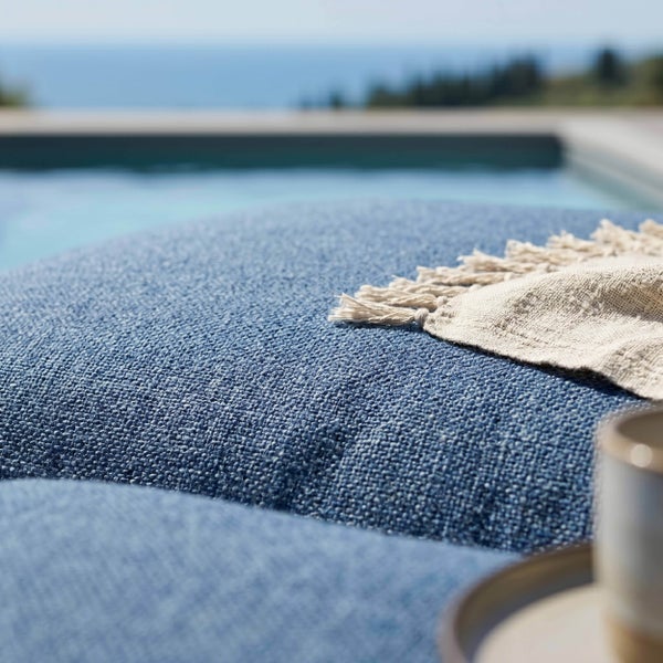 Nahaufnahme eines blauen Outdoor-Kissens mit grober Struktur und einem hellen Fransentuch an einem Pool.