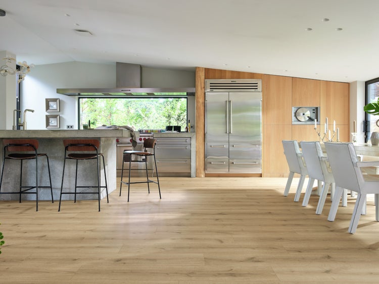 Moderne Wohnküche mit hellem Holzoptik Laminatboden, Kücheninsel aus Beton, Edelstahlgeräten und großem Essbereich mit weißen Stühlen.