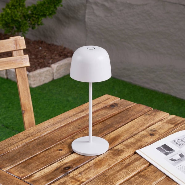 Weiße moderne Akku-Tischleuchte auf einem hölzernen Gartentisch im Außenbereich mit grünem Rasen im Hintergrund.