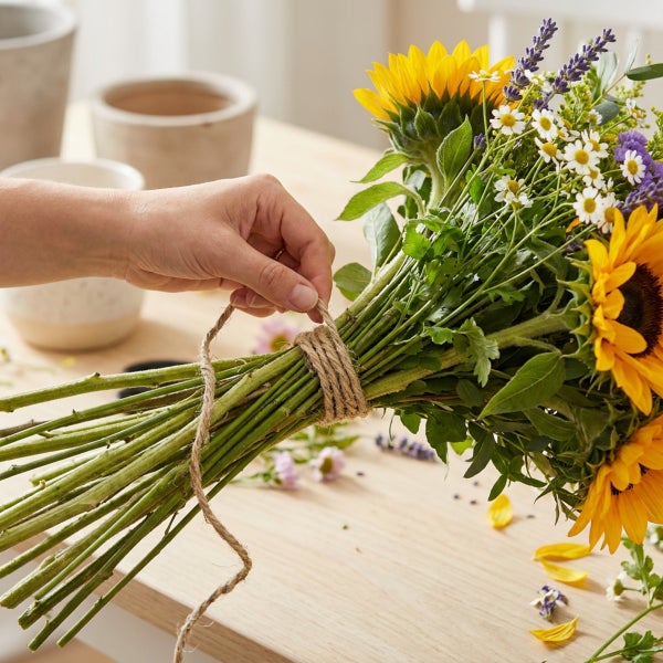 Hand bindet einen Blumenstrauß aus Sonnenblumen, Kamille und Lavendel mit Jutekordel auf einem Holztisch.