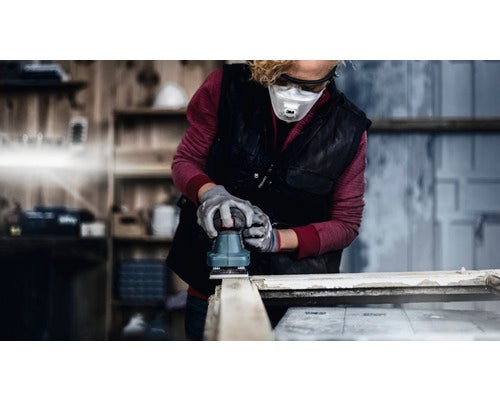 Eine Person schleift Holz mit einem Schwingschleifer. Sie trägt eine Schutzmaske und Schutzbrille.